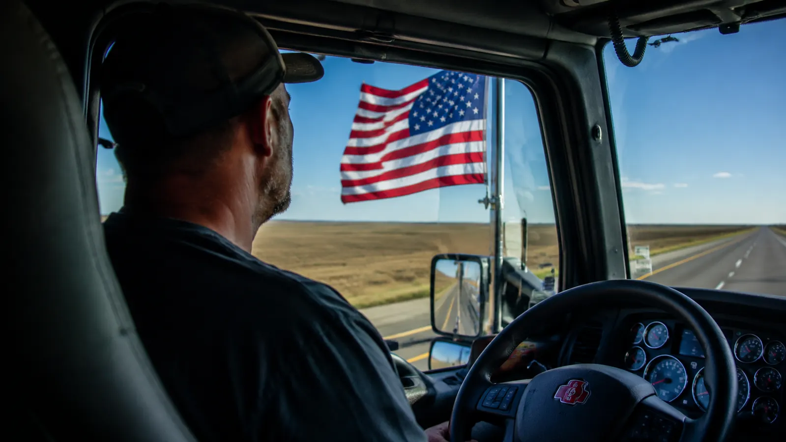 Trucker in a cab looking out at his American flag flying from the truck mirror mount, seen from behind