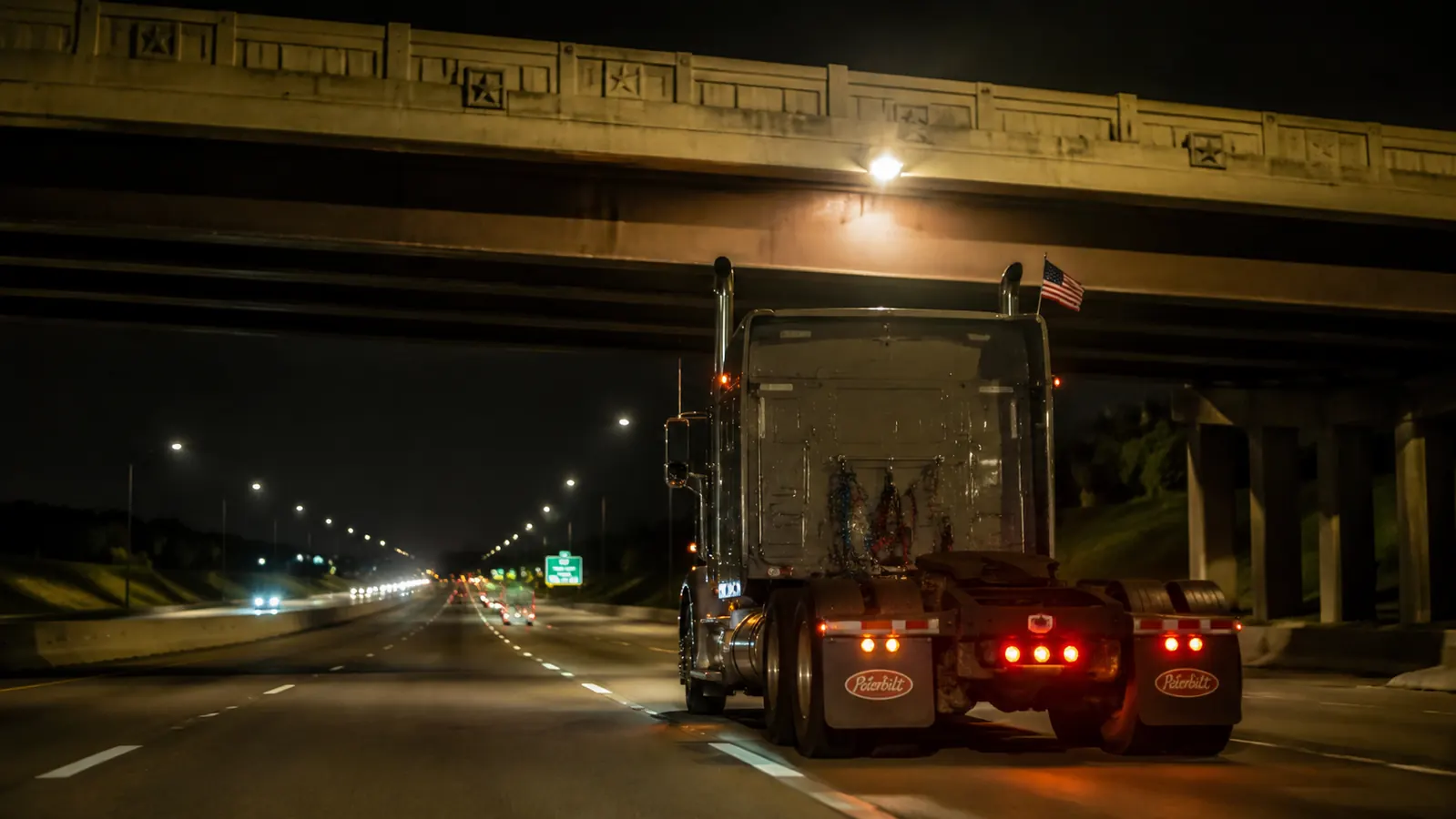 Highway overpass at night with a semi truck passing under, American flag still flying under the lights