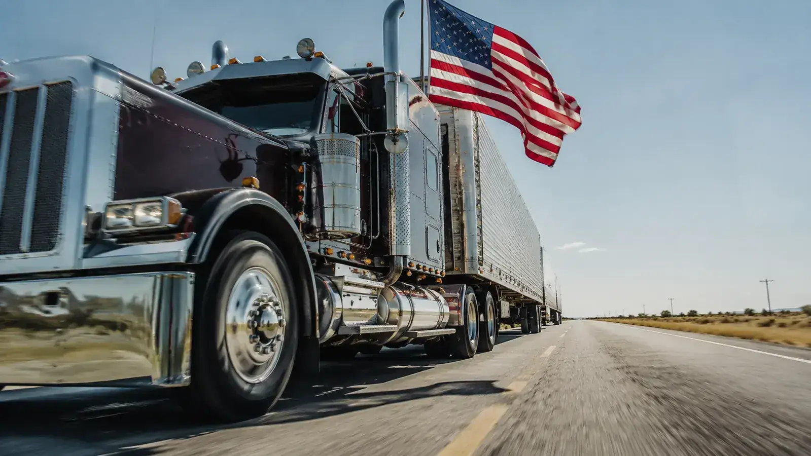 View from alongside a moving semi truck on the highway, American flag visible flying on a pole mounted to the truck, long straight road ahead