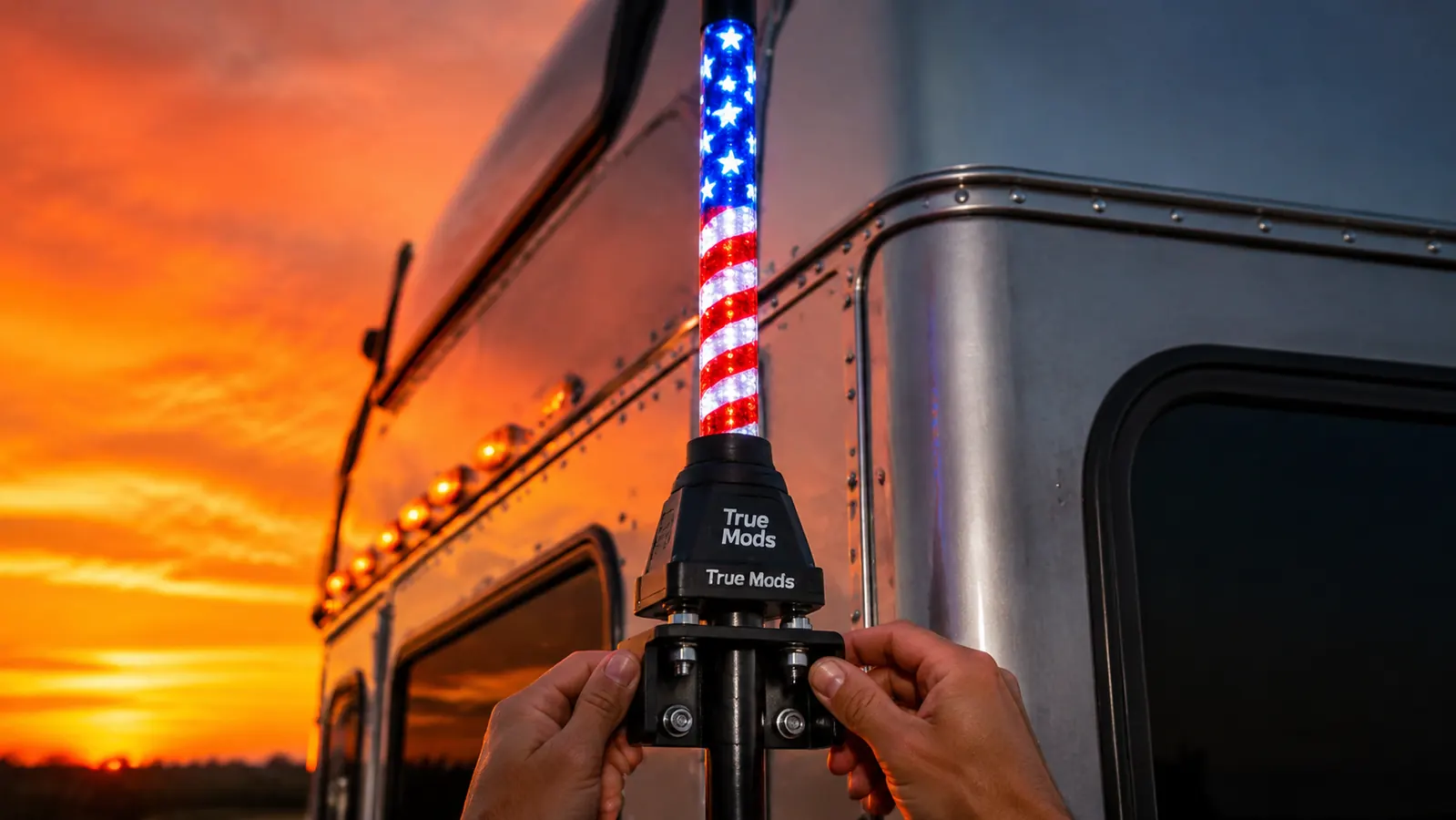 True Mods LED whip light mounted on a semi truck cab-roof bracket, American flag attached and fully unfurled, glowing red at dusk with highway in background